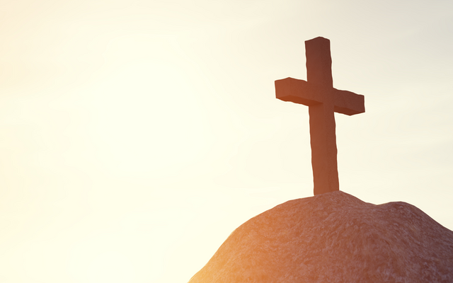 Ein Kreuz steht auf einem Hügel, beleuchtet von warmem Sonnenlicht am Horizont. @Canva